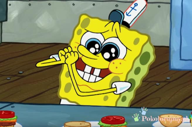 SpongeBob z wielkimi oczami i uśmiechem, trzymający patyk, na tle kuchni.